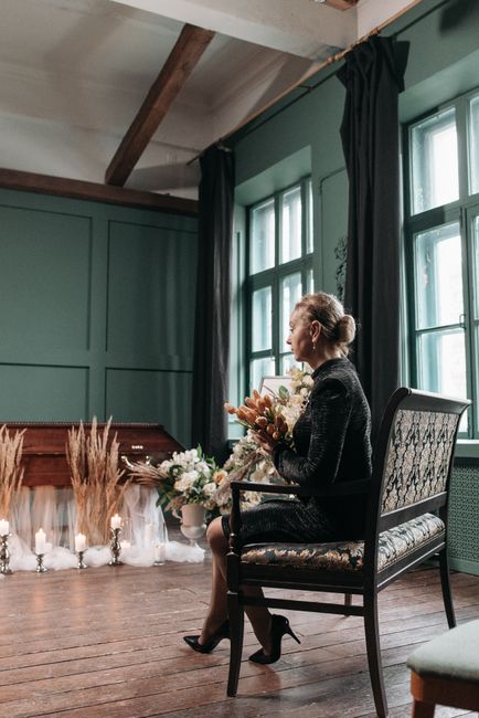 A woman seated on an ornate black wooden chair with upholstered floral fabric, holding a floral arrangement of cream, peach, and soft pink flowers. She is dressed in a black, textured dress, and has h
