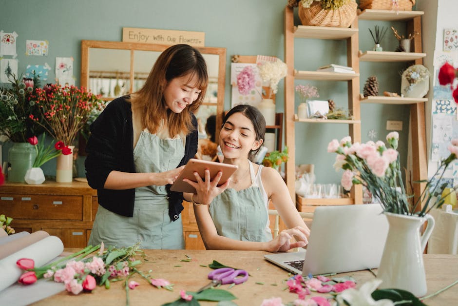 Two women, one standing and one seated, are in a flower shop with a pastel green wall in the background. The standing woman holds a tablet and is showing something to the seated woman, both smiling. O