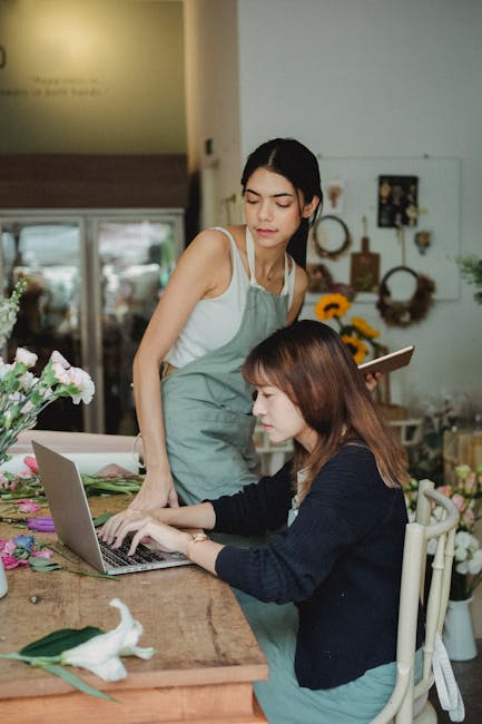 A floral arrangement workshop at Balham Florist, showcasing two women working together. One woman, in a white top and light apron, stands beside a table, observing the other woman who is seated and ty