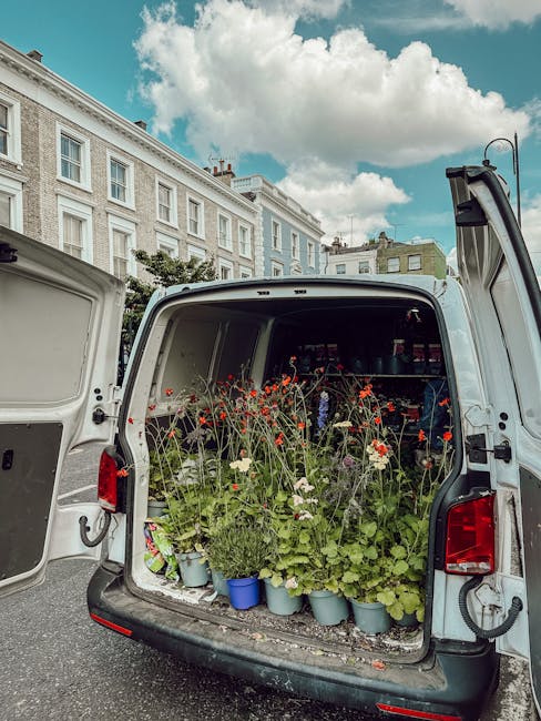 The image shows the open back of a white delivery van parked on a street in a residential area with Victorian-style terrace houses in the background under a partly cloudy sky. Inside the van, there is