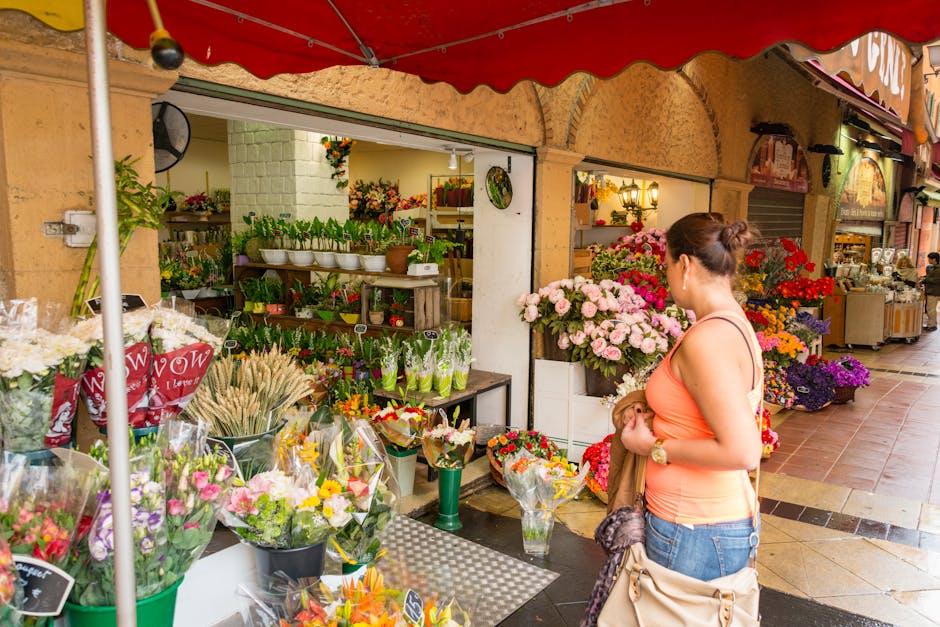 A woman with brown hair tied in a bun, wearing an orange sleeveless top and blue jeans, stands in front of a flower shop at Balham station. She is holding a brown jacket and beige handbag, looking at 