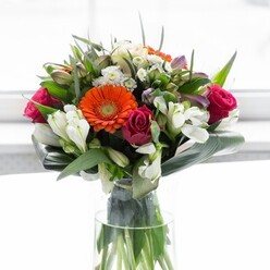 This exquisite Balham bouquet is a masterclass in colour harmony, texture, and local flair. Creamy peach gerbera daisies form a soft halo around bold orange counterparts, their warmth echoing sunlit mornings in the area's tree-lined avenues. Fuchsia-magenta roses contribute an opulent richness, while pristine white alstroemeria and petite spray roses introduce delicate, intricate forms. Glancing between the blooms, subtle pinks and small white fillers add further visual intrigue, making the arrangement feel both abundant and thoughtfully composed. At its base, broad, glossy aspidistra leaves wrap the stems in an elegant embrace. This verdant collar gives the bouquet its full, rounded shape-much like the harmony of Balham's bustling weekends and the calm of its green spaces. All the while, slender green stems gleam through the clear glass vase, reinforcing the display's freshness. Bathed in gentle light, each petal and leaf glows with renewed colour. The bouquet's gentle fragrance-a marriage of rose sweetness and fresh greenery-suggests festive brunches, friendly gatherings, and moments of new beginning. Whether destined for a home on Hayes Grove or celebrating a milestone, this arrangement speaks of optimism, sophistication, and the artisan touch of a local florist.